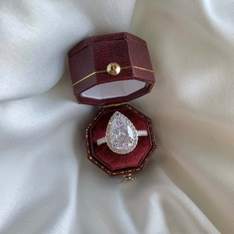 A close-up of a pear-shaped diamond engagement ring with a sparkling halo, set in a silver band. The ring is displayed inside a maroon velvet ring box, resting on a white satin cloth.
