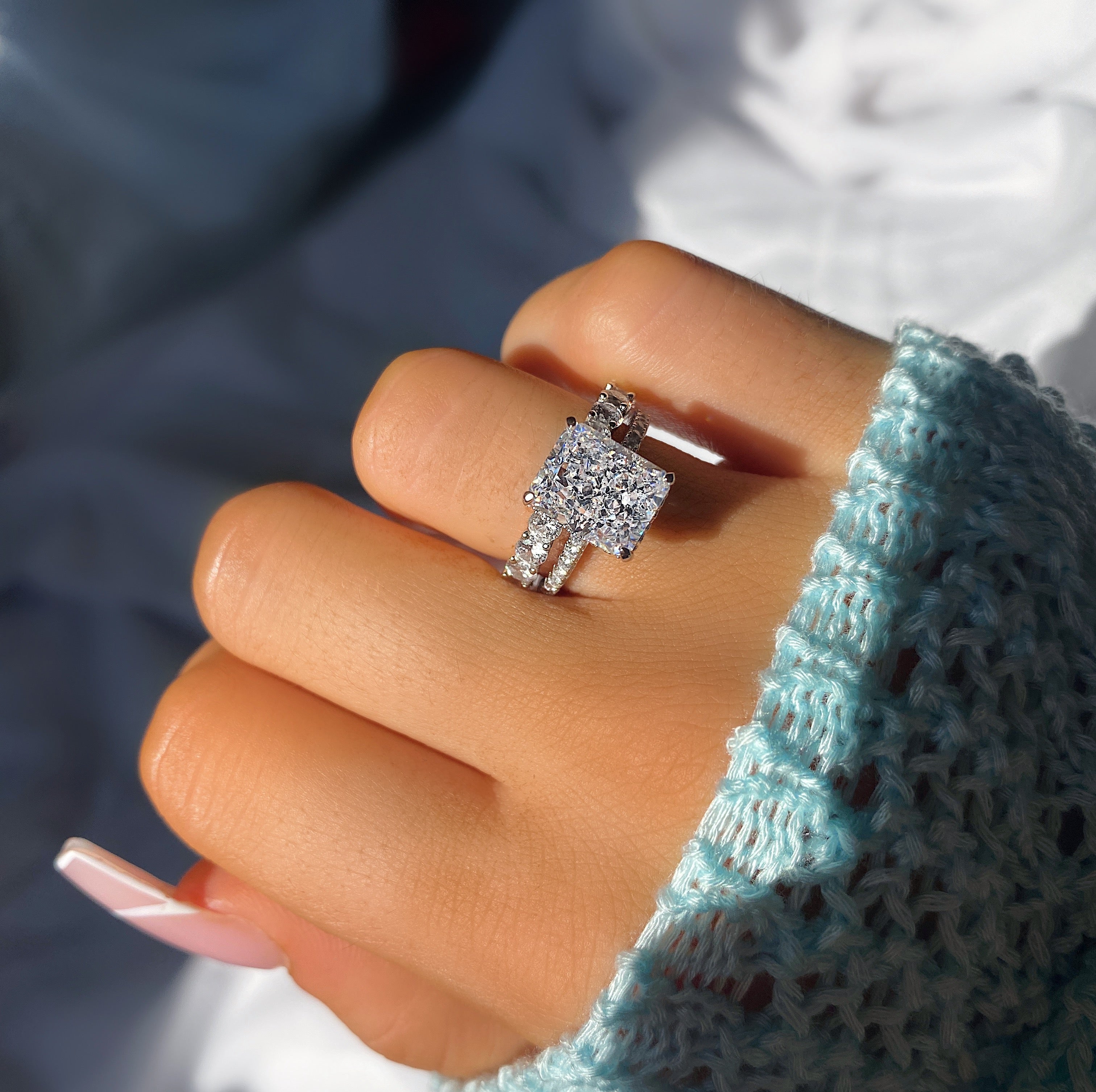 A silver ring with a large rectangular cubic zirconia stone surrounded by a smaller halo of stones, worn on a model wearing  a blue knit jumper.